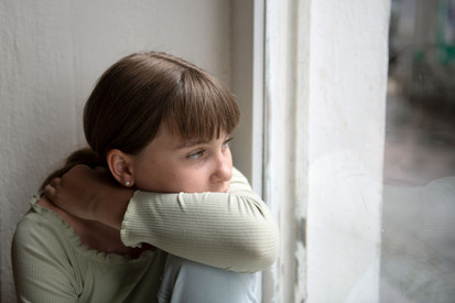 Eine Mädchen sitzt am Fenster und Blickt traurig nach draußen.