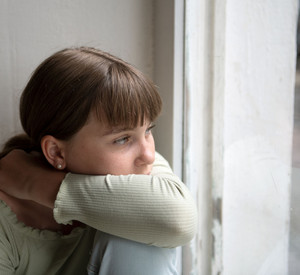 Eine Mädchen sitzt am Fenster und Blickt traurig nach draußen.